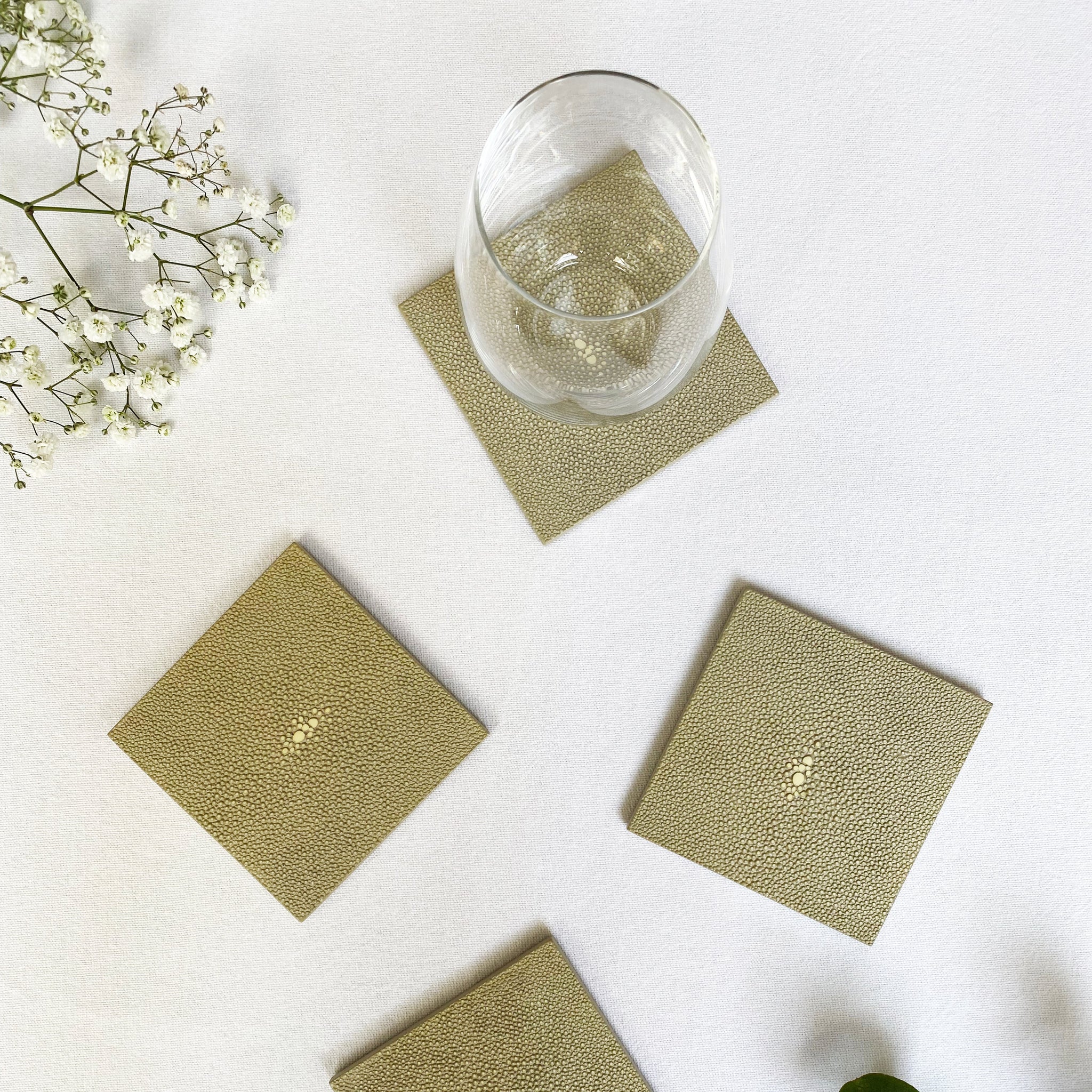 Set of four green coasters on a white surface with a glass of water and decorative plants.
