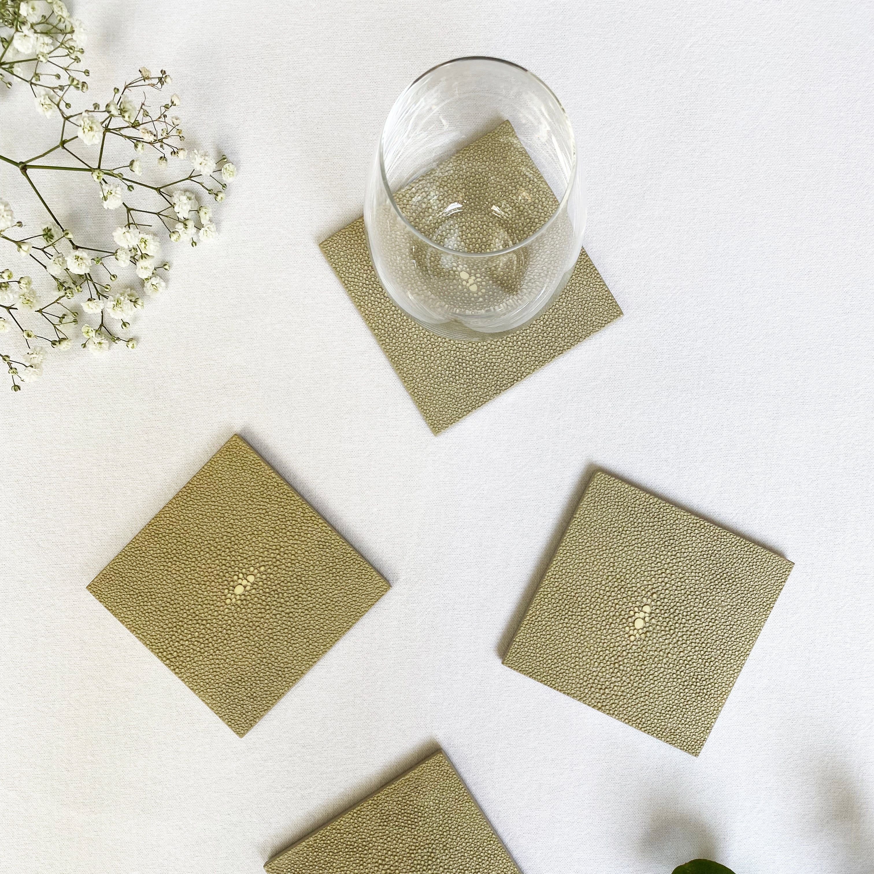 Set of four green coasters on a white surface with a glass of water and decorative plants.