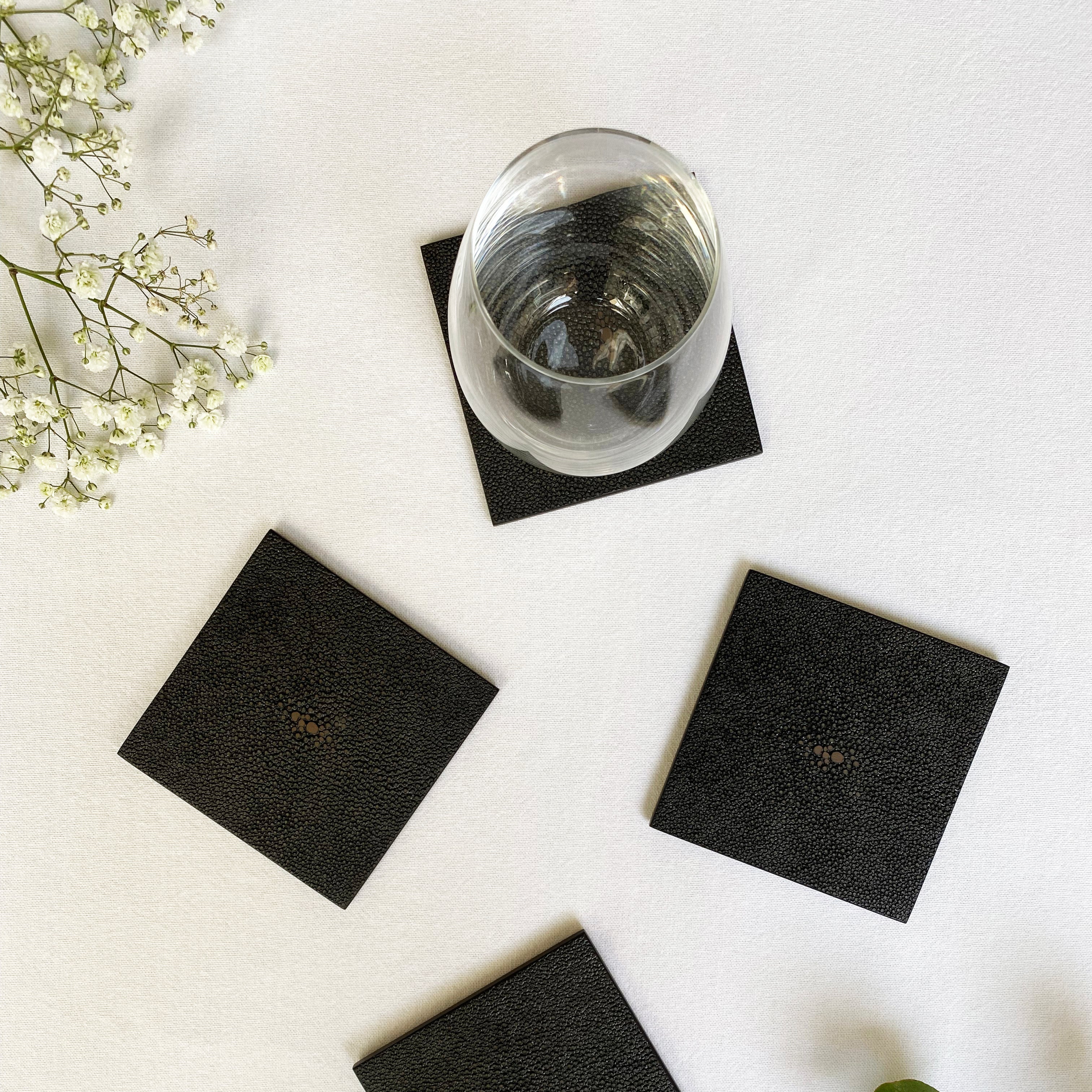 Black square coasters on a white surface with a glass of water and greenery.