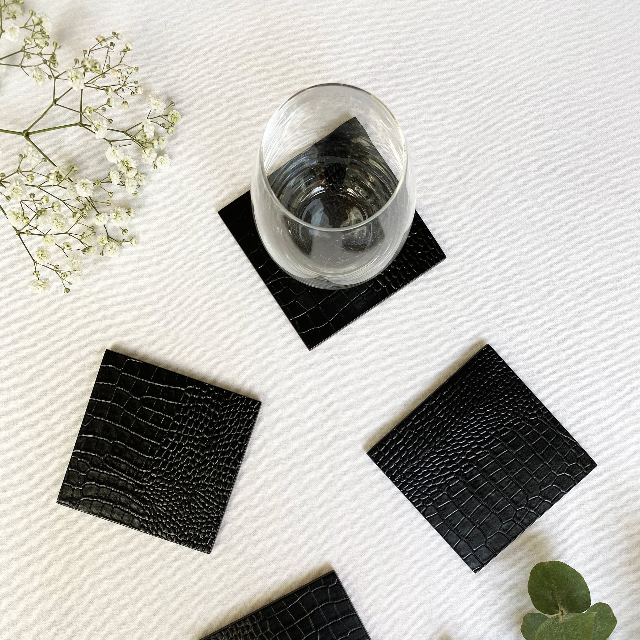 Four black crocodile-patterned coasters on a white surface with a glass of water and greenery.