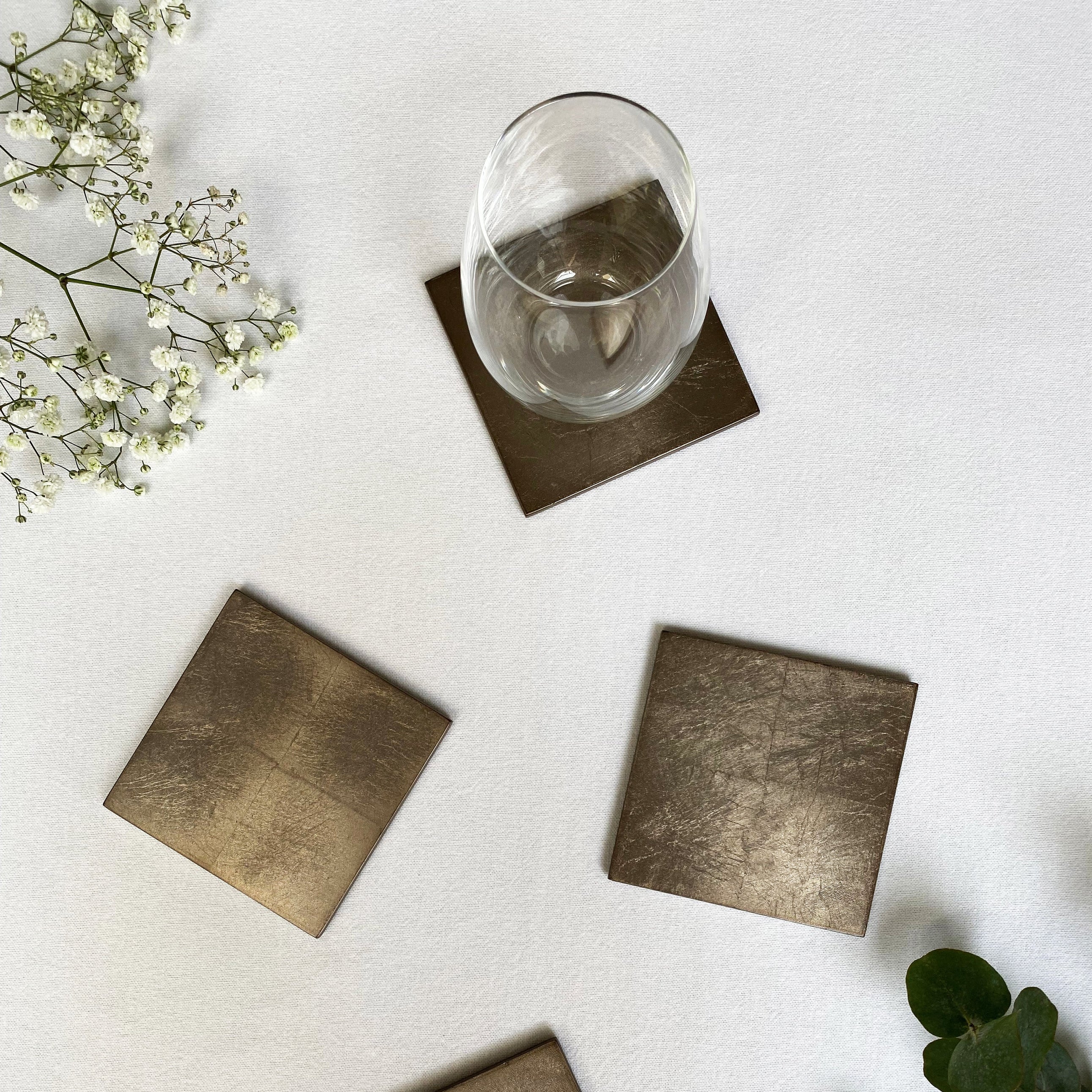 Set of square metallic coasters on a white surface with a glass of water and flowers.