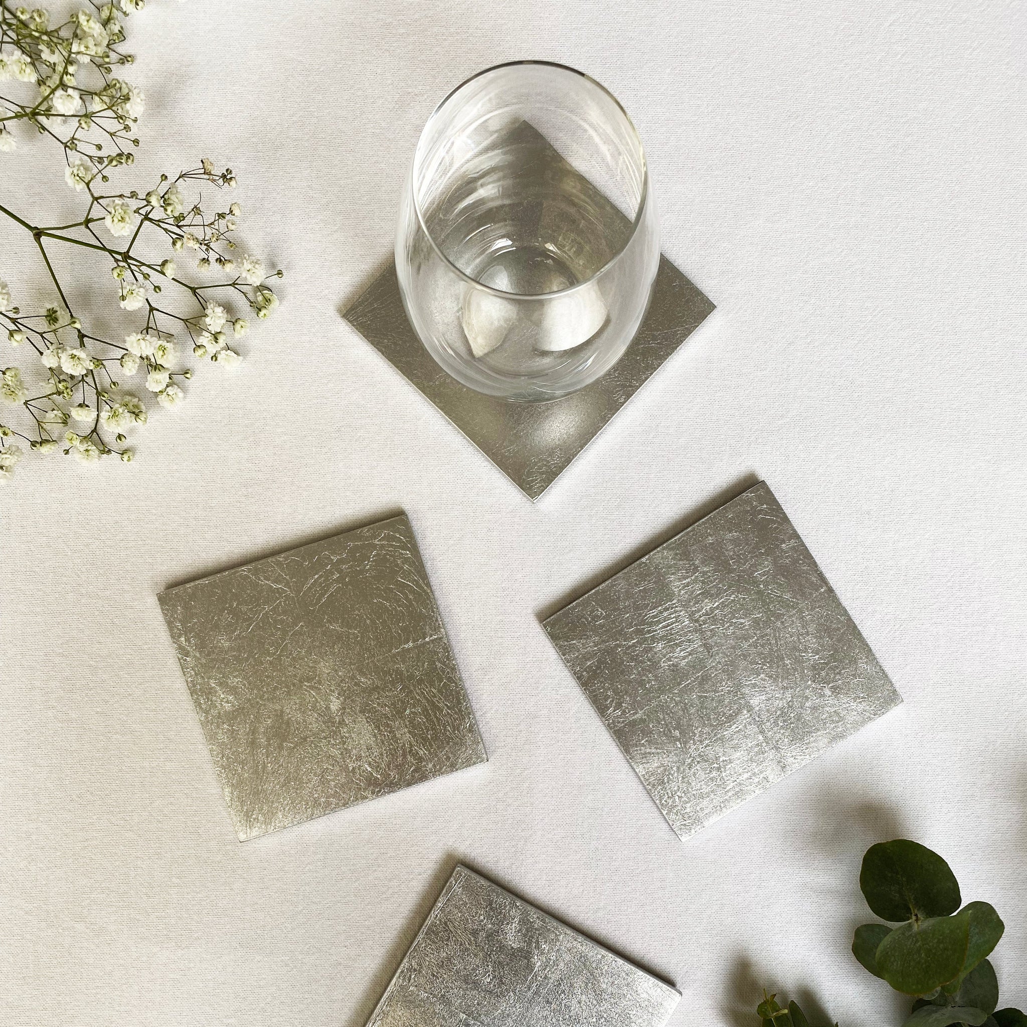 Silver square coasters on a white surface with a glass of water and greenery.