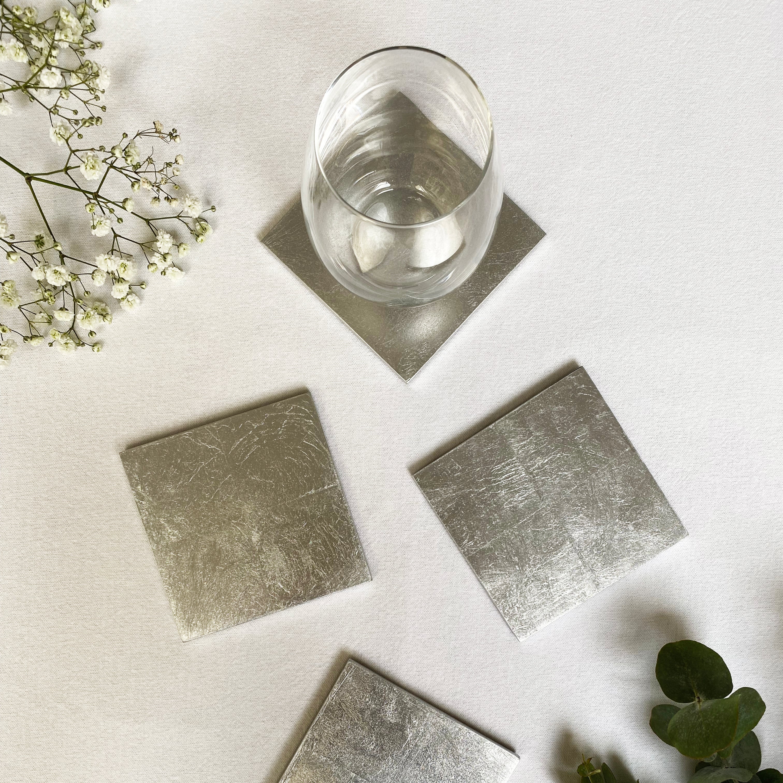Silver square coasters on a white surface with a glass of water and greenery.