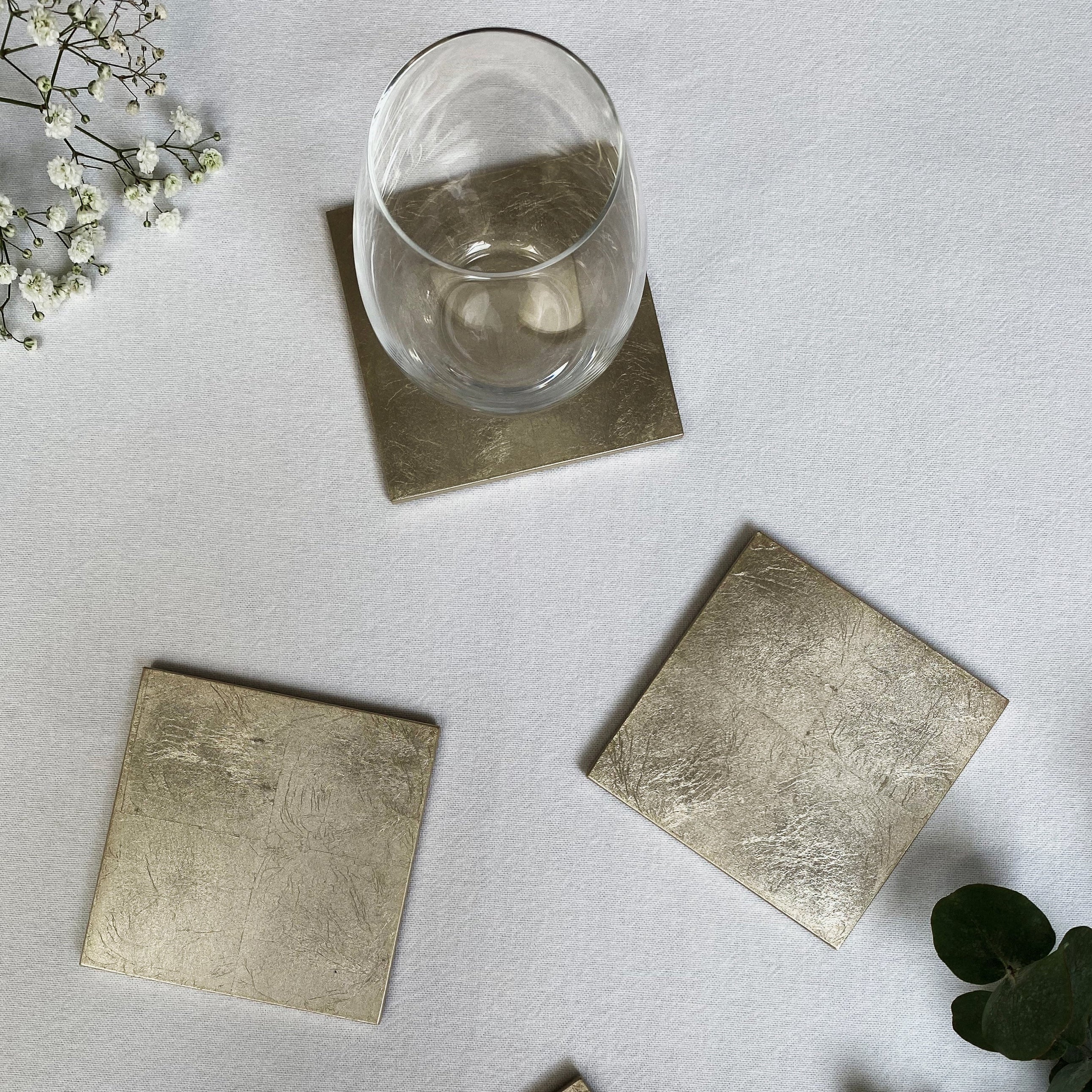 Set of square metallic coasters on a light surface with a glass and greenery.