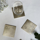 Set of square metallic coasters on a light surface with a glass and greenery.