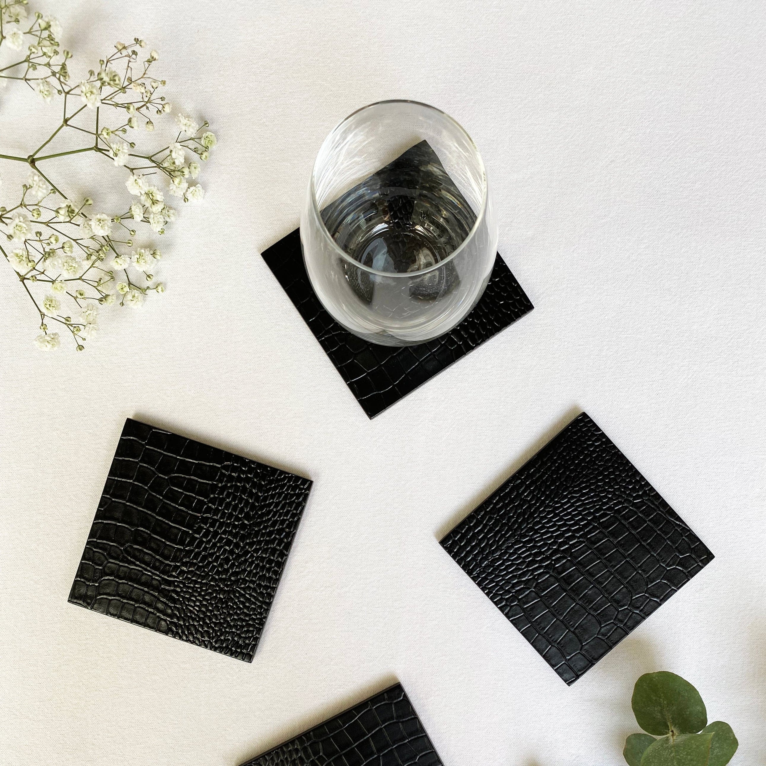 Four black crocodile-patterned coasters on a white surface with a glass of water and greenery.