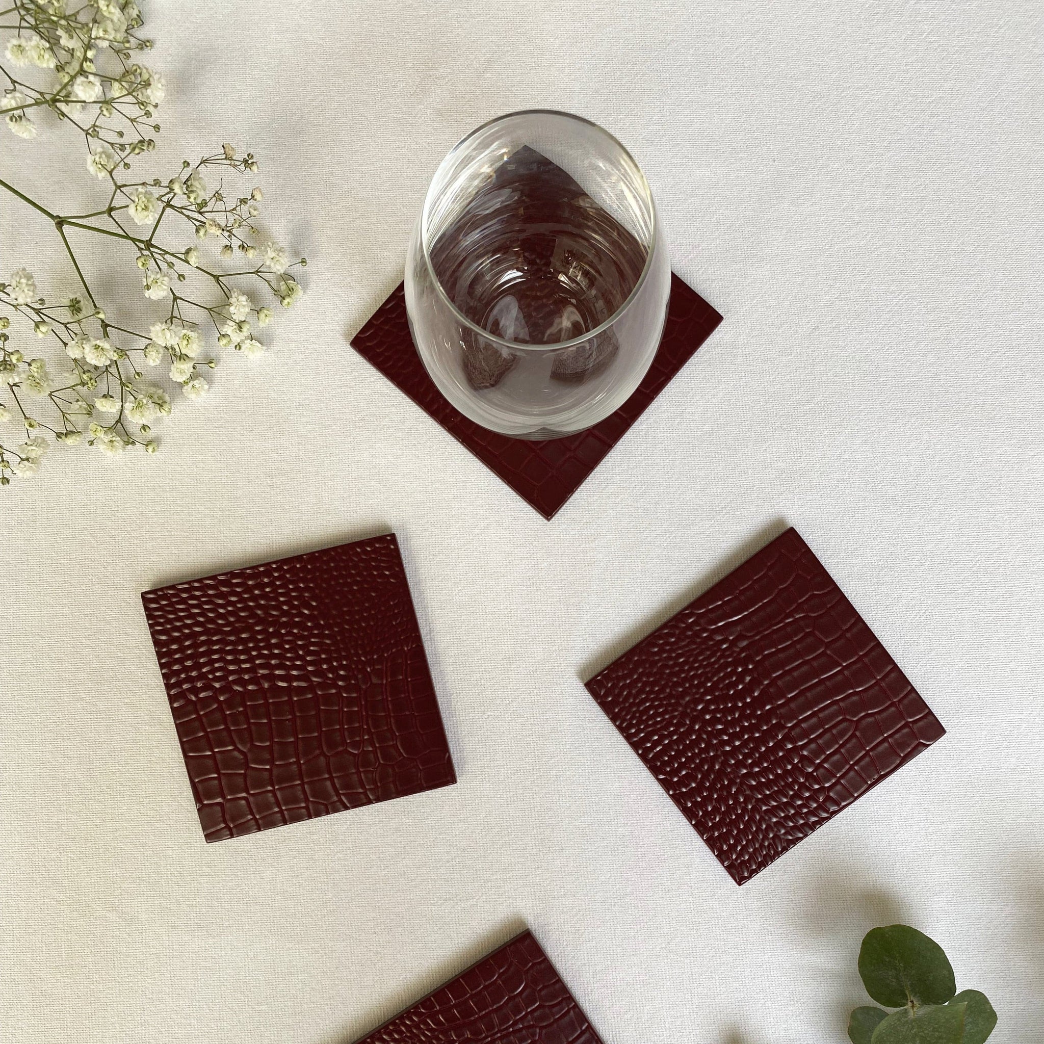 Set of four brown crocodile-textured coasters on a light surface with a glass of water and flowers.